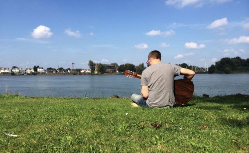 Rear view of man plying guitar while sitting on field by lake