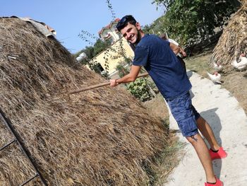 Portrait of man holding gardening fork while standing on footpath against hay