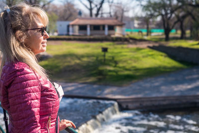 Close side view of a cheerful adult woman with sunglasses standing in the park holding on handrail