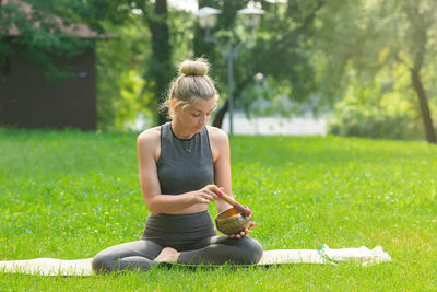 A beautiful woman sitting on the green grass , holds a meditation bowl, drives a stick on it.