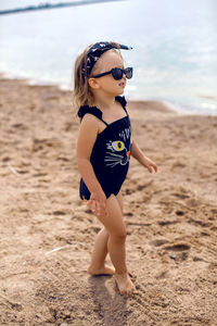 A little girl stands on the shore of the beach in a black bathing suit and black glasses