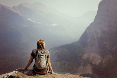 Rear view of woman sitting at cliff against valley and mountains