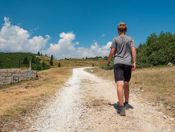 Young blond hair man in shorts and grey t shirt walk on gravel road at beautiful hills. male hiker