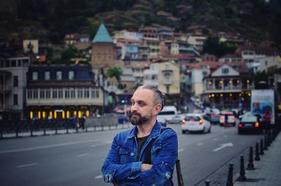 Mid adult man with arms crossed looking away while standing on street in city during sunset