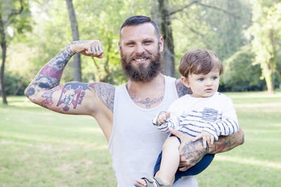 Portrait of smiling bearded man flexing muscle while holding son on field 