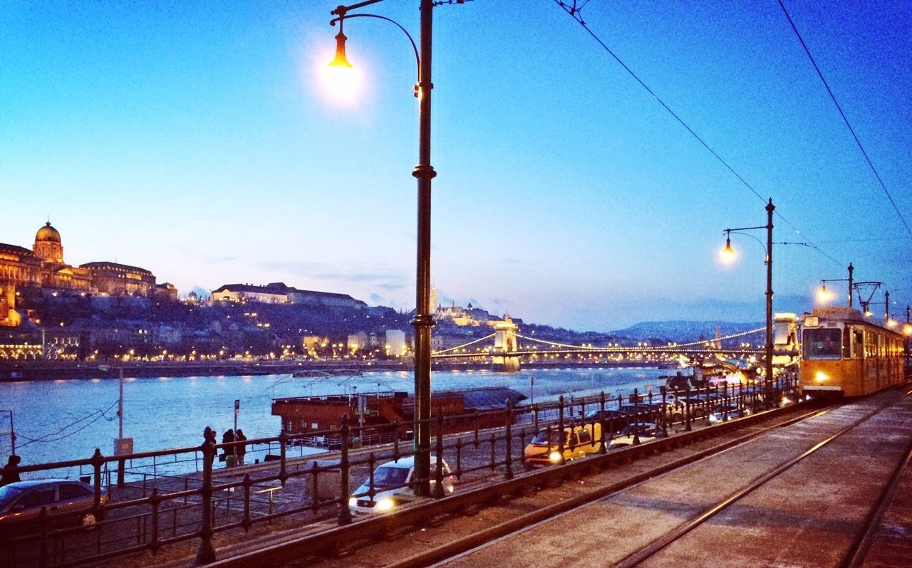 street light, illuminated, lighting equipment, transportation, water, blue, sky, clear sky, railing, mountain, night, dusk, built structure, sea, mode of transport, outdoors, architecture, electricity, lamp post, river