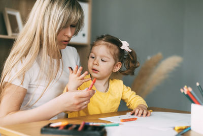 Happy mother and daughter draw together with colored pencils at the table in the room. time together