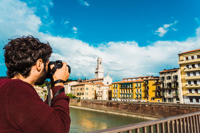 Man photographing by river against sky in city