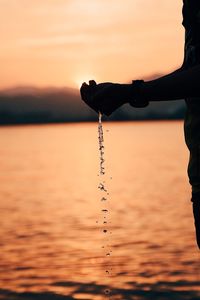 Silhouette person hands holding water over sea against sky during sunset