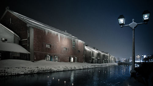 Illuminated street by buildings against sky at night