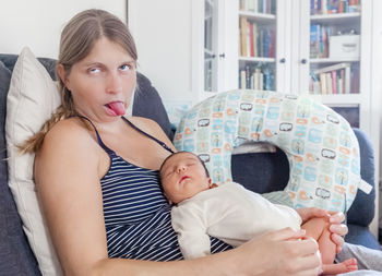 Mother sticking out tongue while sitting with sleeping daughter on sofa at home