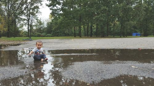 Portrait of cute boy sitting in puddle on road against trees