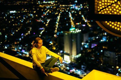 Woman sitting on retaining wall against illuminated city at night