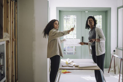 Side view of female design professional explaining blueprint to client at under construction apartment