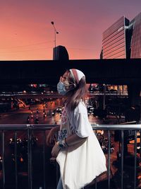 Rear view of young woman standing against buildings during sunset