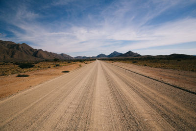 Empty dirt road by mountains against sky