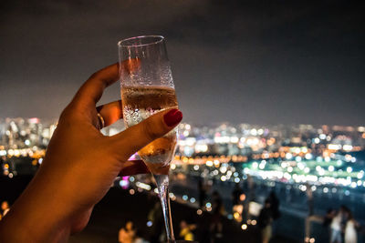 Close-up of hand holding wine glass against illuminated city at night