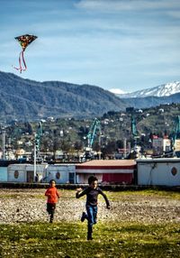 Boys flying kite on field in town against sky
