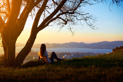 Woman relaxing on grass by tree against sky during sunset