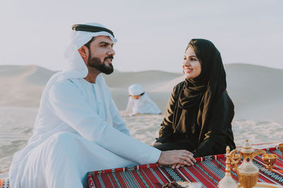 Smiling couple with tea sitting on carpet against son playing at desert