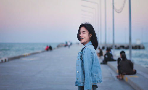 Side view portrait of cheerful young woman standing on promenade against sky during sunset