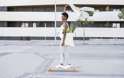 Businesswoman holding blazer while skateboarding with bag of vegetables on footpath