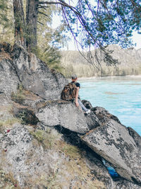 Full length rear view of woman sitting on rock by river in forest