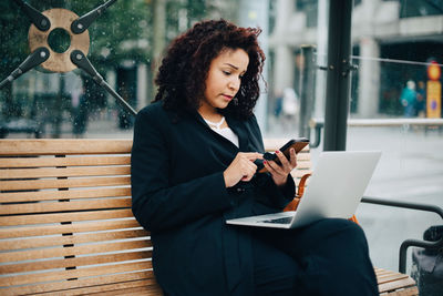 Young woman using mobile phone while sitting on laptop