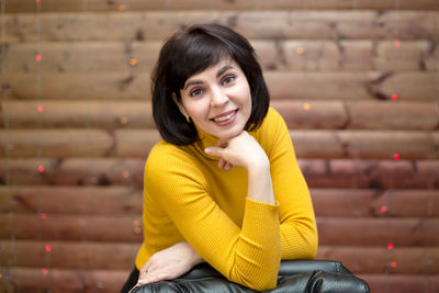 A brunette woman in a yellow jacket with a collar is leaning on a high leather chair.