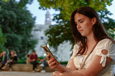 Side view of beautiful woman sitting in park in city, holding mobile phone