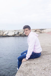 Portrait of young man sitting on retaining wall by sea against clear sky