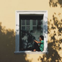 Full length of man sitting on window sill outside house during sunny day