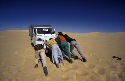 Men and women pushing jeep at desert against clear blue sky