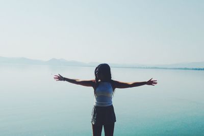 Woman with arms outstretched standing by lake against clear sky