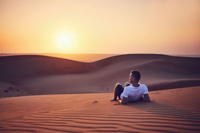 Man leaning on sand dune in desert against sky during sunset