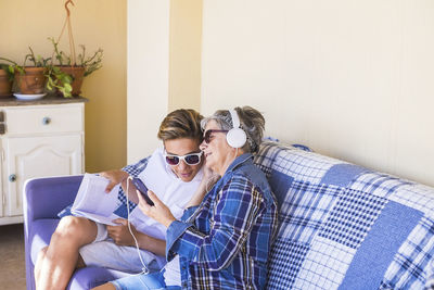 Smiling senior woman listening music through headphones by grandson sitting on sofa at home