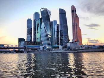 Modern buildings by river against sky during sunset