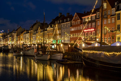 Sailboats moored on illuminated canal by buildings in city at night