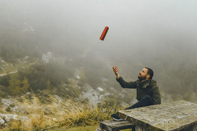 Young man throwing bottle while sitting at table against landscape