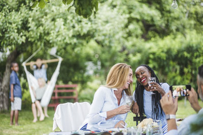 Man with mobile phone photographing smiling women having wine in garden party at weekend