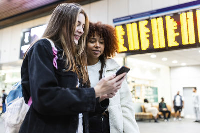 Smiling woman with friend using smart phone at railroad station