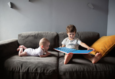 Boy reads a book sitting on the sofa and his brother is lying nearby