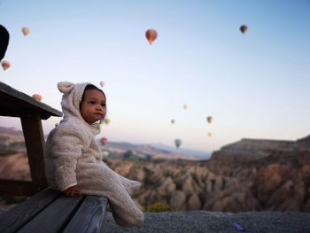 Baby girl sitting on bench against hot air balloons flying over mountains