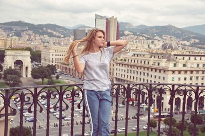 Teenage girl with hand in hair leaning on railing against buildings in city