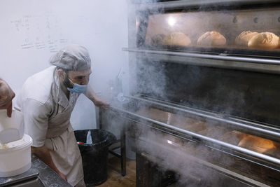 Male baker checking breads in oven while baking in kitchen at bakery