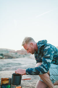 Blonde camper with scruff and camo jacket boils water for dinner