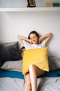 Delighted female sitting on soft bed with cushion and enjoying weekend with closed eyes at home