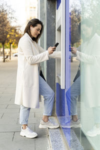 Stylish female entrepreneur using smart phone while withdrawing money from atm