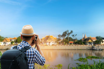 Rear view of mature man photographing while standing by river against clear sky