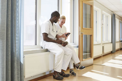 Full length of male and female nurses using smart phone while sitting on window sill at hospital corridor